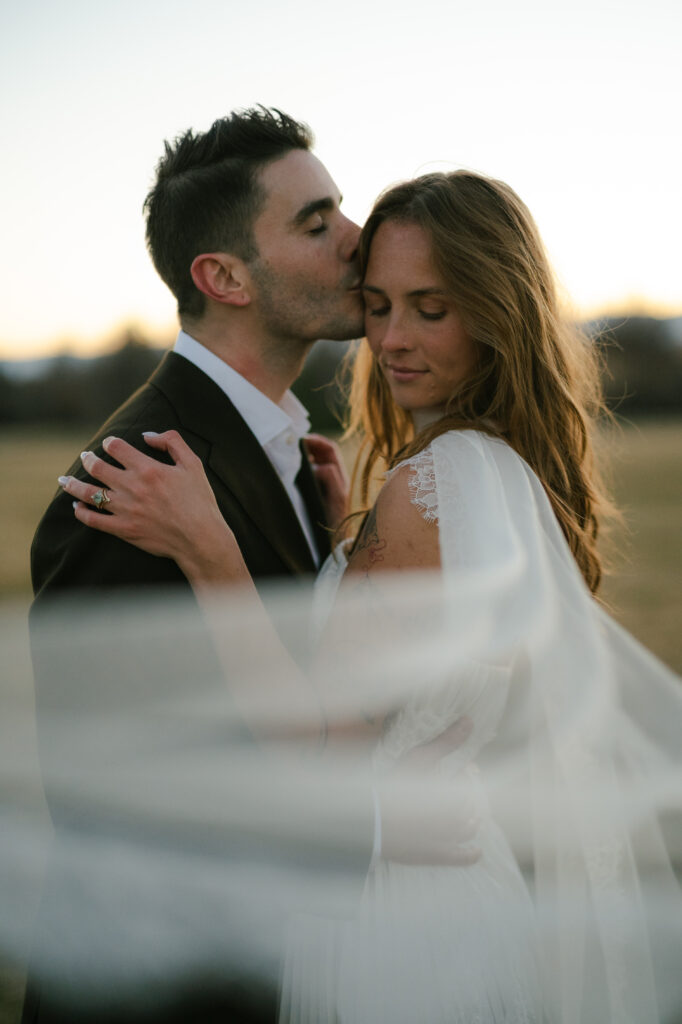 Groom kissing bride's temple with flowy fabric in the wind